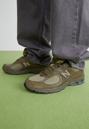 Brązowe buty sportowe New Balance noszone z luźnymi szarymi jeansami, stojące na zielonym dywanowym podłożu.
