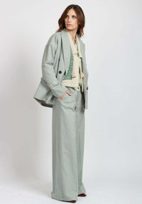 Donna che indossa un completo oversize grigio chiaro con pantaloni a gamba larga e una sciarpa in maglia beige sopra una maglietta a fantasia verde, in piedi contro uno sfondo semplice.