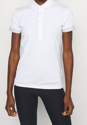 Persoon draagt een nauwsluitend wit poloshirt met korte mouwen en knoopsluiting en een zwarte broek, staand voor een effen achtergrond.