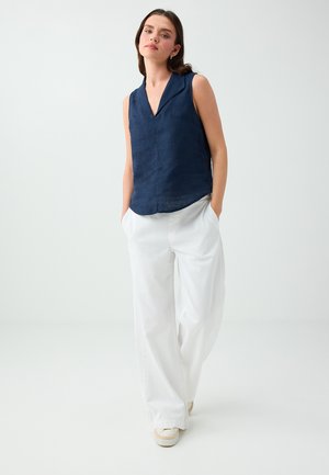 Frau mit dunklen Haaren, die eine ärmellose marineblaue Bluse, weiße weite Hose und weiße Sneakers trägt, steht mit den Händen in den Taschen vor einem hellen Hintergrund.