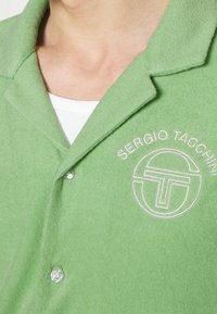 Polo de manga corta en verde, confeccionado en tejido suave, con un logo bordado en blanco y un collar clásico con cierre de botones.