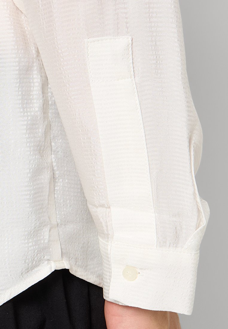 Blusa blanca texturizada con un sutil patrón a rayas, que presenta un puño con botón y un acabado de cuello limpio y estructurado.