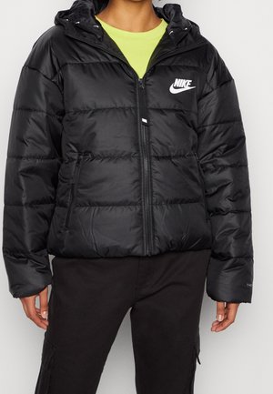 Osoba nosząca czarną pikowaną kurtkę Nike z kapturem, zapinaną na zamek, na limonkowej koszulce i czarnych spodniach.