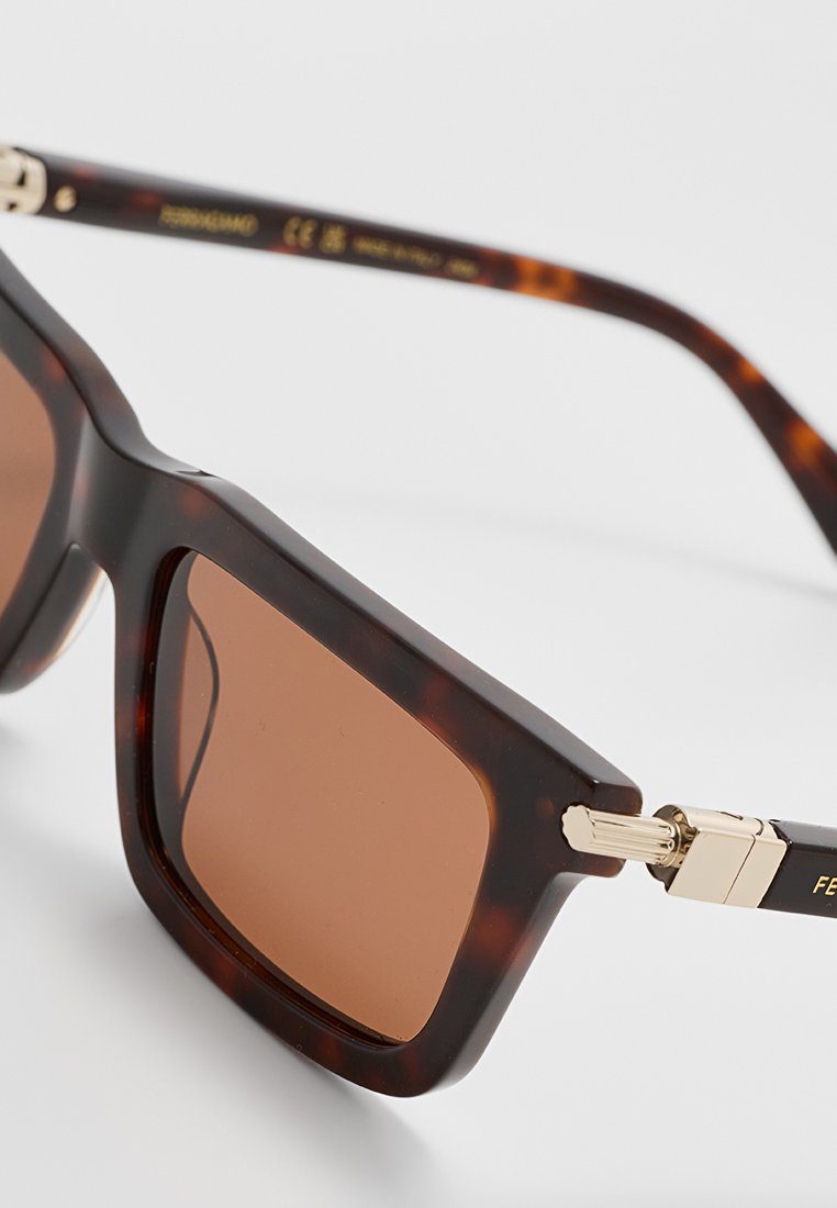 Lunettes de soleil en écaille de tortue brune avec des verres carrés et une monture épaisse. Accents dorés sur les branches et verres teintés bruns.