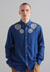 Blaues Jeanshemd mit langen Ärmeln und Kragen, das auf der Brust mit kunstvollen weißen gestickten Blumenmustern verziert ist.