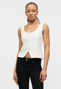 Γυναίκα με πλεγμένα μαλλιά, φοράει άσπρο αμάνικο πουκάμισο με κουμπιά και μαύρο παντελόνι, στέκεται μπροστά σε απλό ανοιχτό φόντο.