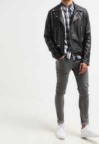 Černá kožená motorkářská bunda se stříbrným kováním, vrstvená přes modrou a bílou kostkovanou košili. Šedé oprýskané skinny džíny a bílé tenisky.
