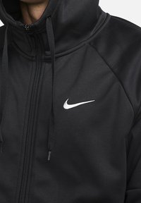 Sudadera con cremallera negra y capucha ajustable, fabricada en tejido suave, que presenta un logo blanco de Nike en el pecho.