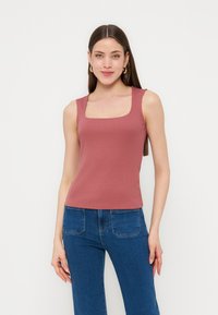 Γυναίκα με μακριά καστανά μαλλιά φοράει χρυσούς κρίκους στα αυτιά, ροζ τοπ χωρίς μανίκια με τετράγωνο ντεκολτέ και ψηλόμεσο μπλε τζιν, στέκεται μπροστά σε λευκό φόντο.