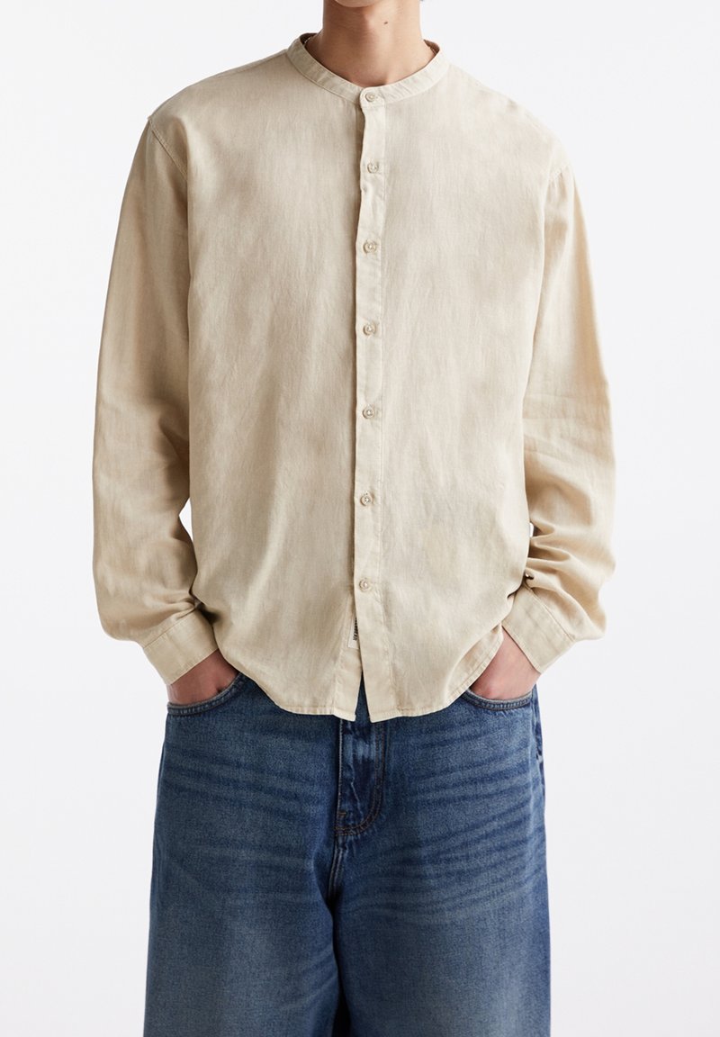 Personne portant une chemise beige boutonnée avec un col mao et des manches longues, associée à un jean bleu, mains dans les poches, sur fond blanc.
