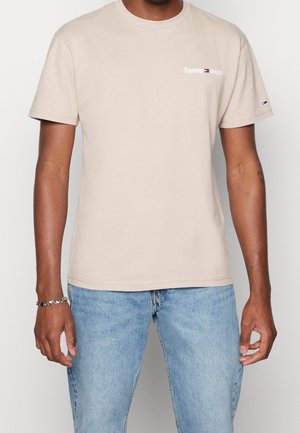 Mężczyzna w beżowej koszulce Tommy Jeans i jasnoniebieskich dżinsach, stojący na tle jednolitego białego tła, z ramionami swobodnie opuszczonymi wzdłuż ciała.