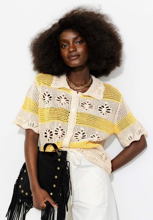 Femme aux cheveux bouclés portant un haut en crochet rayé jaune et crème, un pantalon blanc, un collier en chaîne dorée, tenant un sac à main noir à franges.