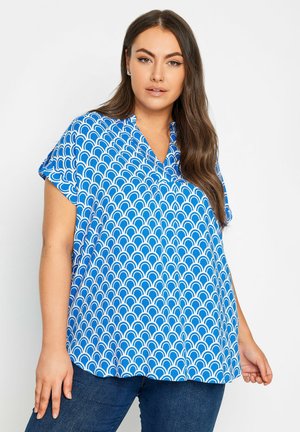 Blusa azul de manga corta con un patrón blanco de conchas repetido, con un corte relajado y diseño de escote en V. Se lleva con jeans de mezclilla oscuros.