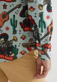 Chemise à manches courtes en tissu bleu clair avec des motifs contrastants colorés de cactus, de guitares et de notes de musique.