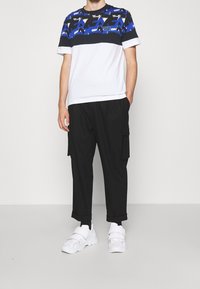 Pánský outfit zahrnující bílou a modrou vzorovanou košili s krátkým rukávem, černé cargo kalhoty s kapsami a bílé tenisky.