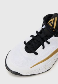 Chaussure de sport blanche avec des lacets noirs, en tissu texturé et un accent doré. Présente une languette en maille noire et une semelle blanche mouchetée.
