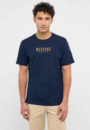 Junger Mann mit lockigem Haar, der ein dunkelblaues T-Shirt mit Mustang-Logo und beige Hosen trägt, vor einem einfarbigen hellen Hintergrund stehend.