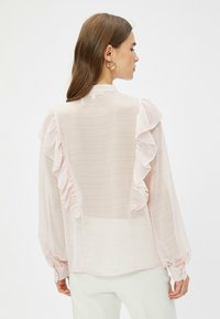 Lys pink skjorte lavet af gennemsigtig stof, med frynse-detaljer langs skuldrene og ryggen, lange ærmer og knapmanchetter.