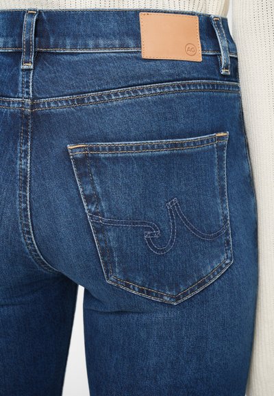 Temno modri denim kavbojke imajo zadnji žep z okrasnim prešitim vzorcem in svetlo rjav usnjen etiket na pasu z logotipom.