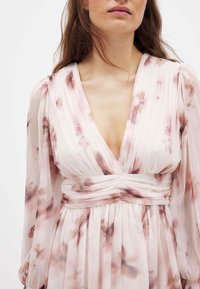 Kvetlična obleka v svetlo rožnati barvi z naborkom in globokim V-izrezom. Dolgi prosojni rokavi in zbrana pasna linija poudarjata silhueto.
