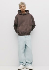 Felpa oversize marrone con tasca a marsupio, abbinata a jeans a gamba larga azzurri e sneakers bianche. Il tessuto ha una consistenza morbida.