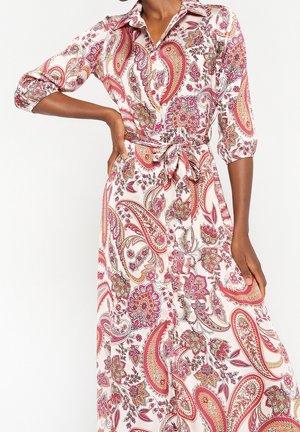 Kvinde iført en hvid langærmet kjole med røde og pink paisley blomsterprint bundet i taljen, poserer med den ene hånd på hoften.