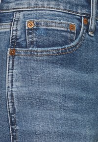 Denimtyg i ljusblått, med en framficka med kobbernitar och synlig sömnad. Sliten textur ger extra detalj.