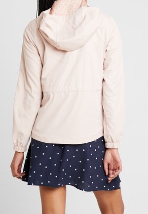 Frau trägt eine hellrosa Kapuzenjacke mit elastischen Bündchen und einen dunkelblauen Rock mit weißen Punkten, von hinten gezeigt.