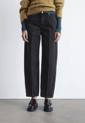 Schwarze weite Jeans aus Denim mit hoher Taille, vorderen Taschen und glatter Textur, kombiniert mit dunklen Slippern und einem gestrickten Pullover.