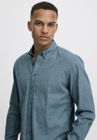 Chemise bleu clair boutonnée en tissu texturé, avec col, manches longues et boutons contrastants le long de la patte de boutonnage.