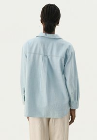 Camicia in denim azzurro chiaro con piega posteriore, maniche lunghe con polsini a bottoni e colletto classico; texture morbida e vestibilità rilassata.
