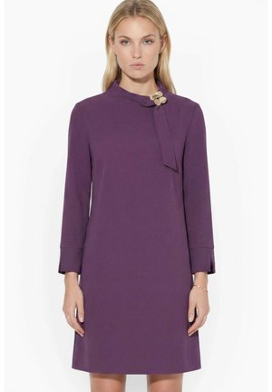 Woman with straight blonde hair wearing a long-sleeve purple knee-length dress with a gold flower brooch on the collar.