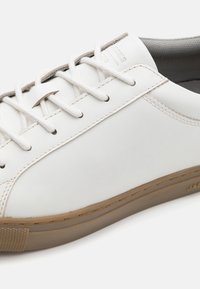 White leather sneaker with round toe, flat laces, and light brown rubber sole. Features subtle stitching and a textured inner lining.