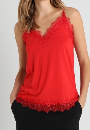 Kobieta w czerwonym, koronkowym topie bez rękawów z dekoltem w serek, w czarnych spodniach, ręce swobodnie w kieszeniach.