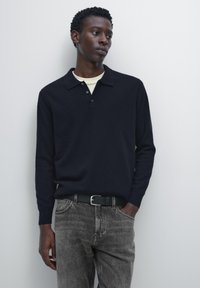 Navy knitted pullover with a collar, three buttons, and long sleeves, paired with gray denim jeans and a black belt. Plain background.