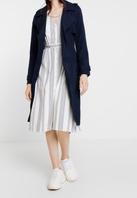 Marinblå lång trenchcoat över en vit klänning med vertikala ränder; modellen bär vita sneakers, jackan har en dubbelknäppt design och bälte.