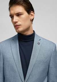 Lichtblauwe gestructureerde blazer met een smalle revers, gecombineerd met een donkerblauwe turtleneck. Bevat een kleine speld op de linkermouw.