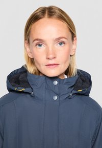 Marineblauwe geïsoleerde jas met een rits- en drukknoopsluiting, een capuchon en zachte voering. Heeft metalen accenten en een geborduurd patroon.