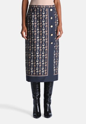 Jupe midi marine avec motif chaîne beige et blanc, cinq grands boutons dorés sur un côté, portée avec des bottes noires texturées montant aux genoux.