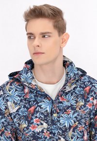 Giacca leggera floreale di colore navy con parroti colorati e foglie tropicali, dotata di zip frontale e cappuccio. Tessuto testurizzato.