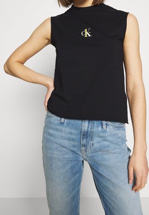Person trägt ein ärmelloses schwarzes Top mit kleinem CK-Logo und hellblaue Jeans, die linke Hand ruht auf der Hüfte vor einfarbigem Hintergrund.