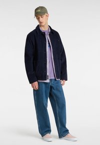 Giacca di velluto a coste blu navy con tasche frontali e chiusura a bottoni, indossata sopra una maglietta lavanda, abbinata a pantaloni di denim blu rilassati e sneakers chiare.