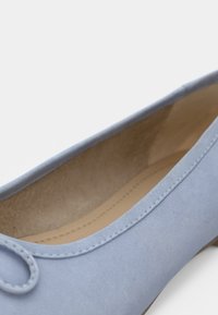 Μπλε σουέντ μοκασίνι με στρογγυλεμένη μύτη και διακοσμητικό λουράκι στο επάνω μέρος. Το εσωτερικό είναι μπεζ, με λεία υφή.