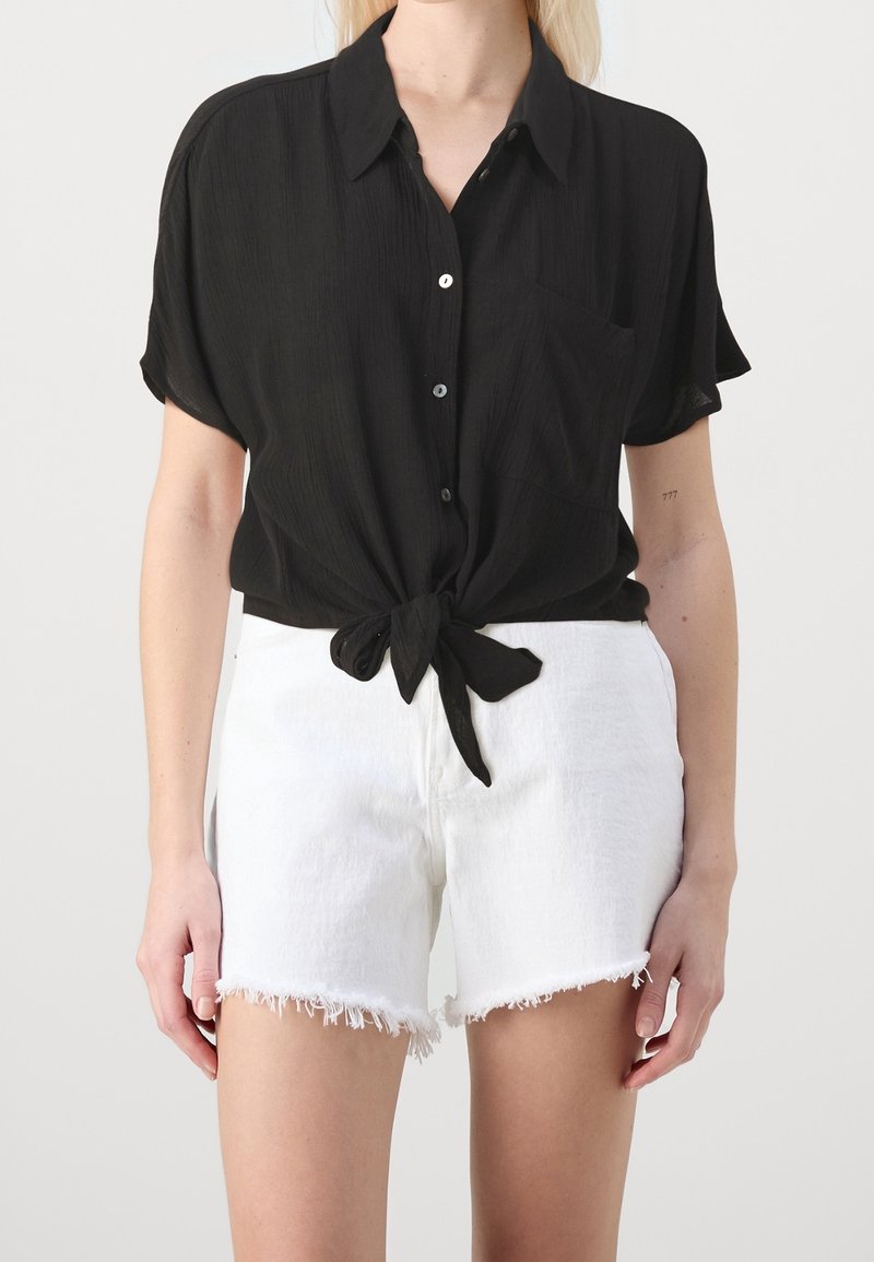 Femme portant une chemise noire à manches courtes boutonnée nouée à la taille et un short en jean blanc effiloché, debout devant un fond uni.
