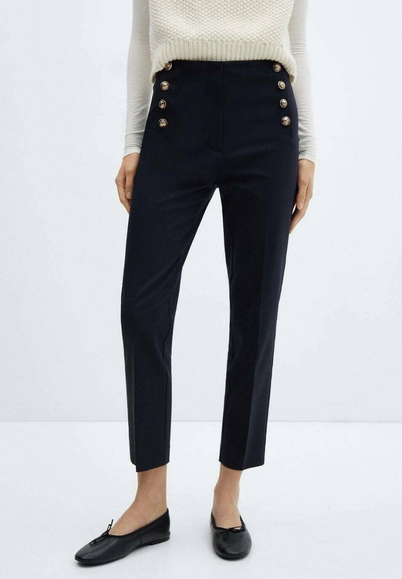 Femme portant un pantalon noir taille haute avec des boutons dorés décoratifs et des ballerines noires, associé à un haut en maille blanc.