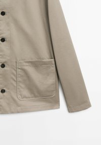 Lichtbeige jas gemaakt van gladde stof, met een knoopsluiting aan de voorkant, lange mouwen en een vierkant zakje aan de linkerkant.