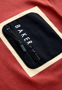 Černá látková kapsa se zipem a bílým nápisem „BAKER by Ted Baker“ na červeném a béžovém pozadí oděvu.
