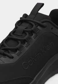 Κοντινό πλάνο μαύρου αθλητικού παπουτσιού Calvin Klein που δείχνει τα κορδόνια με θηλιές, την ταινία για τράβηγμα και το ανάγλυφο όνομα της μάρκας στην πλαϊνή πλευρά.