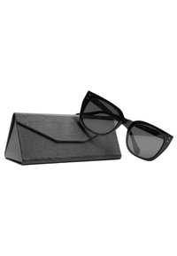 Occhiali da sole oversize neri con lenti scure, forma angolare e bordi appuntiti, esposti accanto a un astuccio triangolare nero con texture.