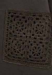 Temno rjav kos oblačila z kvadratnim kvačenim krojačem floralnih vzorcev, obdanim s teksturiranim robom, na gladkem tekstilnem ozadju.